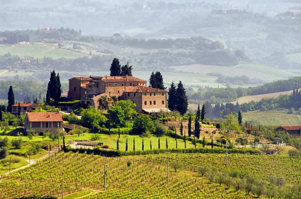 Et charmerende landskab i Toscana, Italien, med en traditionel vingård omgivet af grønne vinmarker og cypresser. Stenhuse med røde tegltage ligger på en bakketop med en betagende udsigt over det bølgende landskab. Baggrunden viser de karakteristiske bløde bakker dækket af vinmarker, oliventræer og skov.