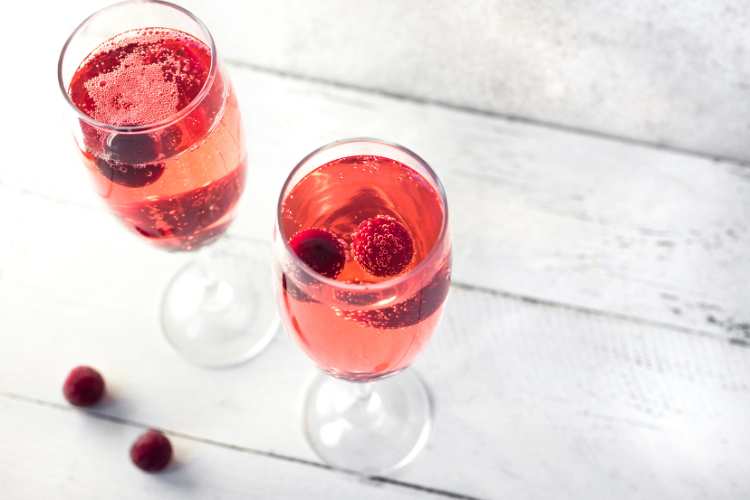 To champagneglas fyldt med den klassiske Kir Royale velkomstdrink, en boblende rosarød cocktail pyntet med friske hindbær. Glassene står på en hvid træoverflade med et par ekstra hindbær ved siden af, hvilket skaber en elegant og festlig stemning.