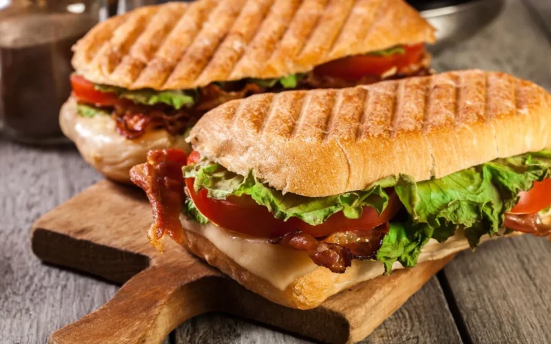 Panini grill med sandwich, tomat, ost og salat på et træskærebræt